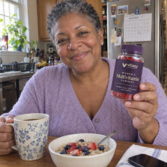 Woman in her late 50s holding a jar of Vitasy Women's Multivitamin Gummies at the breakfast table with coffee and oatmeal