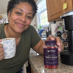 Woman in her 40s holding a jar of Vitasy Women's Multivitamin Gummies and a mug by the coffee maker in her kitchen