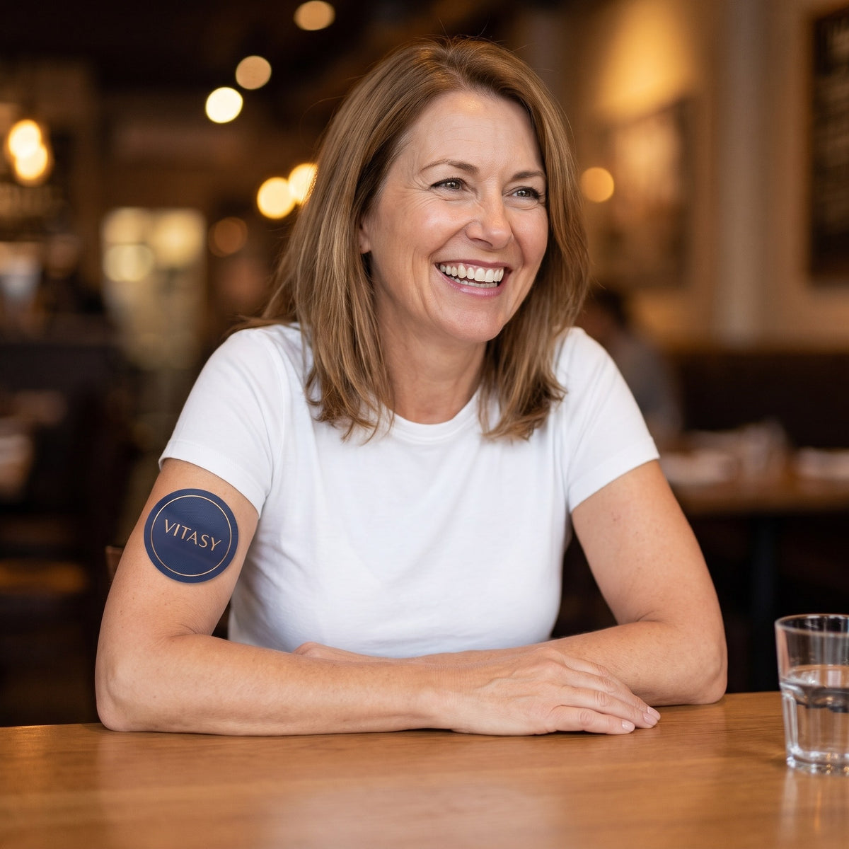 Smiling woman at a restaurant table with a Vitasy patch on her arm