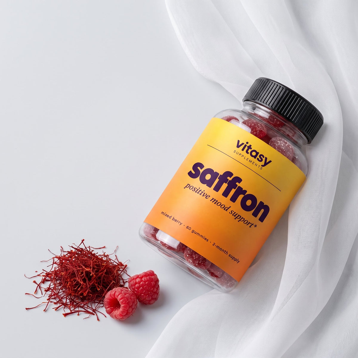Bottle of Vitasy Saffron Gummies lying on sheer fabric with saffron threads and raspberries