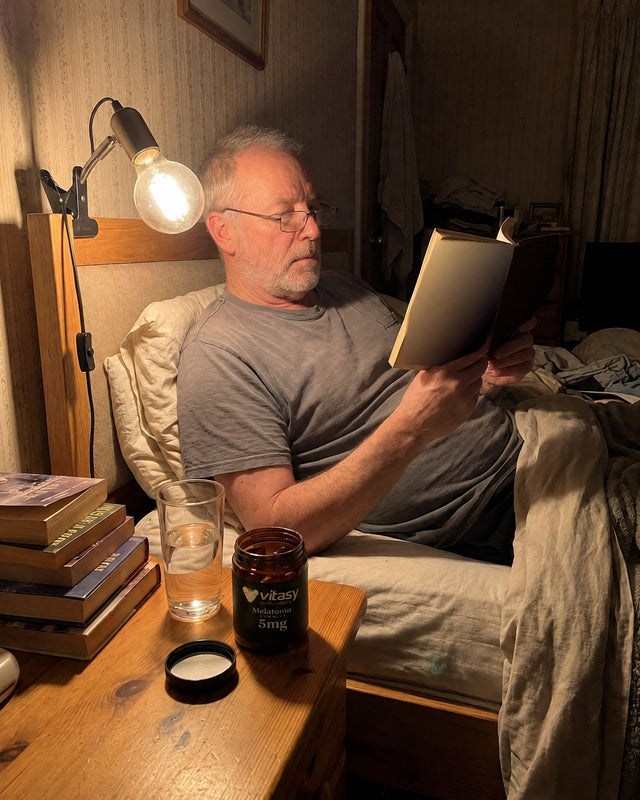 Man reading in bed under a lamp, Vitasy Melatonin Gummies jar on the nightstand