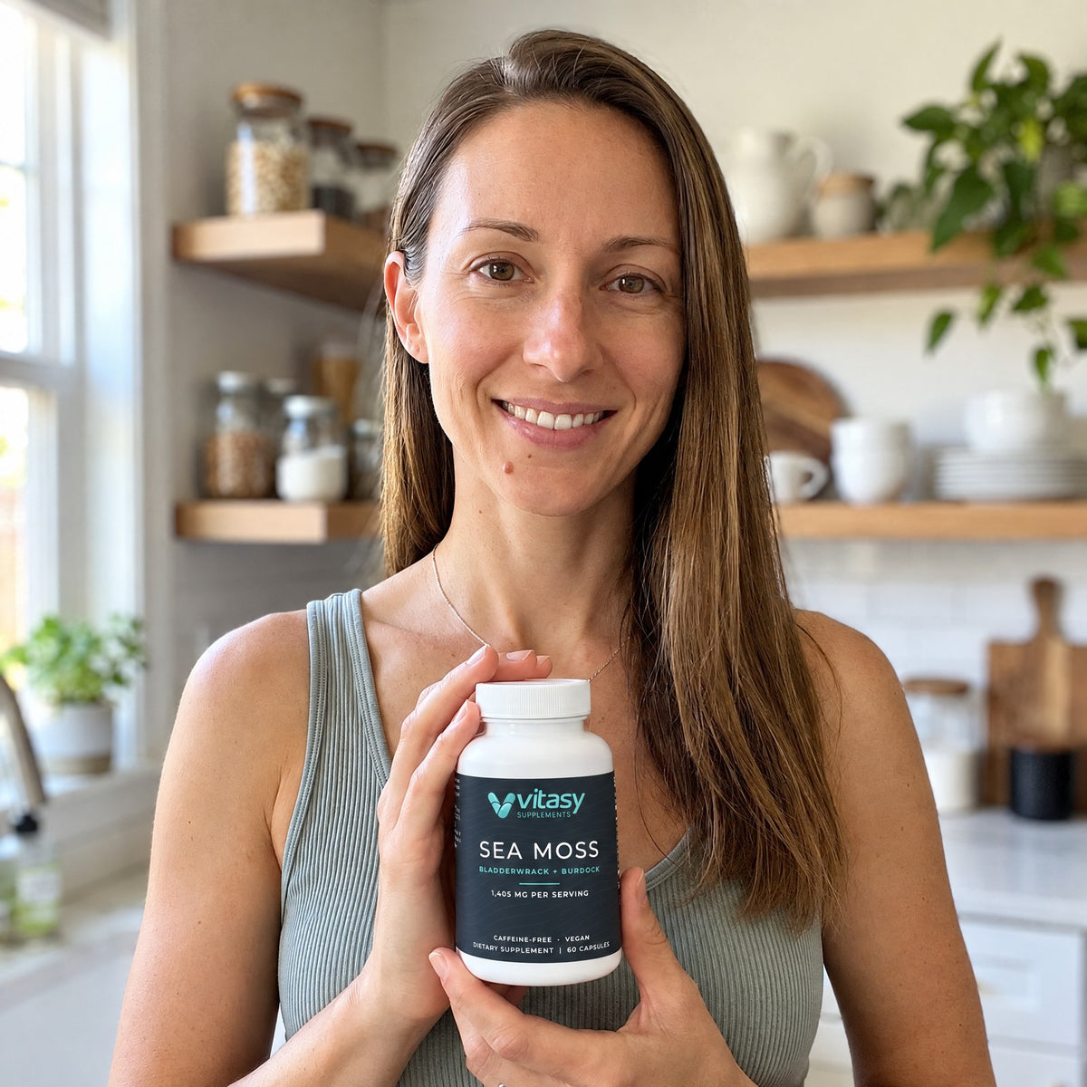 Smiling woman in a bright kitchen holding a bottle of Vitasy Sea Moss Capsules
